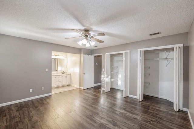 unfurnished bedroom featuring a textured ceiling, ceiling fan, dark wood-type flooring, connected bathroom, and multiple closets