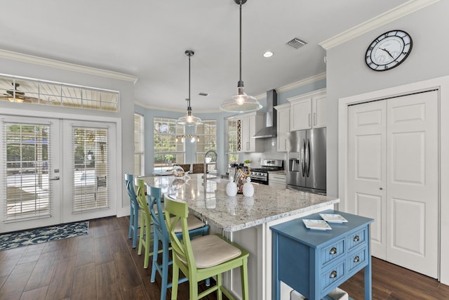 kitchen featuring pendant lighting, wall chimney range hood, appliances with stainless steel finishes, a kitchen island with sink, and white cabinets