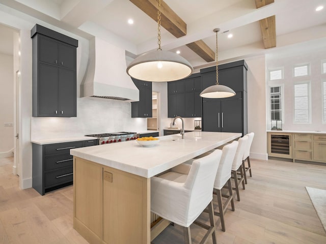 kitchen featuring custom range hood, beverage cooler, a center island with sink, beamed ceiling, and hanging light fixtures