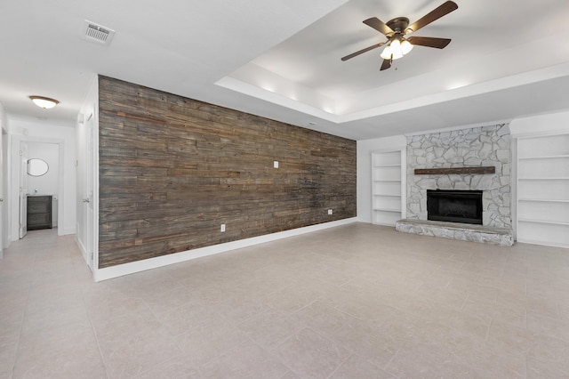 unfurnished living room featuring built in shelves, a tray ceiling, ceiling fan, a stone fireplace, and light tile patterned flooring