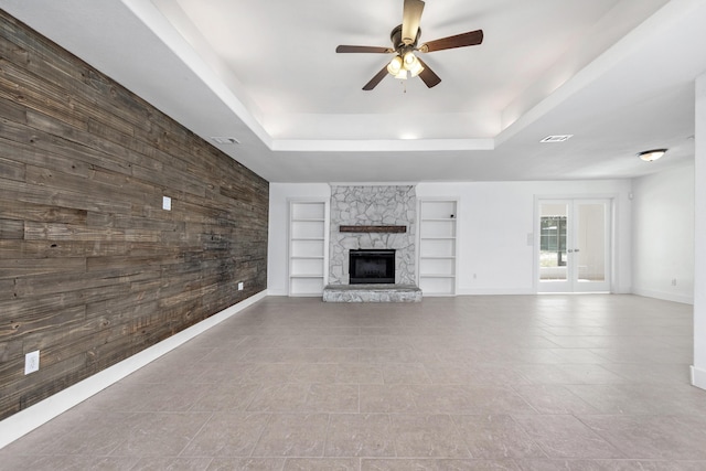 unfurnished living room with a raised ceiling, ceiling fan, a stone fireplace, and built in features