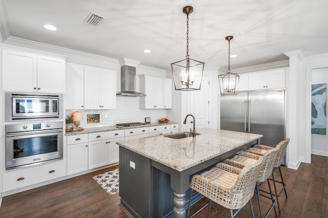 kitchen featuring a kitchen island with sink, wall chimney range hood, sink, built in appliances, and decorative light fixtures