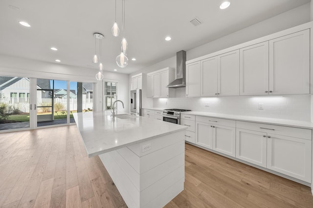 kitchen with a center island with sink, decorative light fixtures, light wood-type flooring, and wall chimney range hood