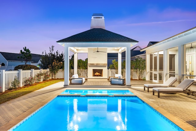 pool at dusk with an outdoor fireplace, a gazebo, ceiling fan, and a patio