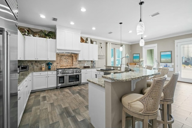 kitchen featuring light stone countertops, high end appliances, a center island with sink, white cabinetry, and hanging light fixtures