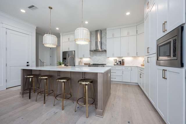 kitchen with appliances with stainless steel finishes, decorative light fixtures, an island with sink, white cabinets, and wall chimney exhaust hood