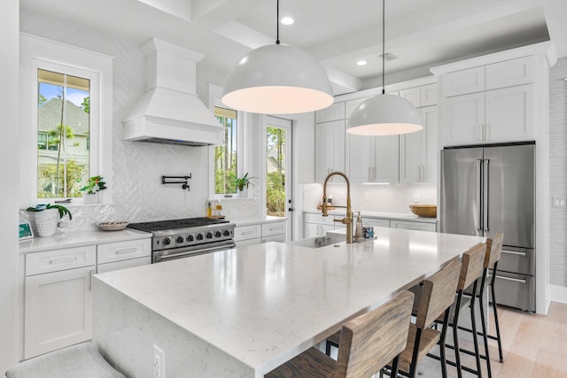 kitchen with decorative light fixtures, white cabinets, high end appliances, custom exhaust hood, and a center island with sink