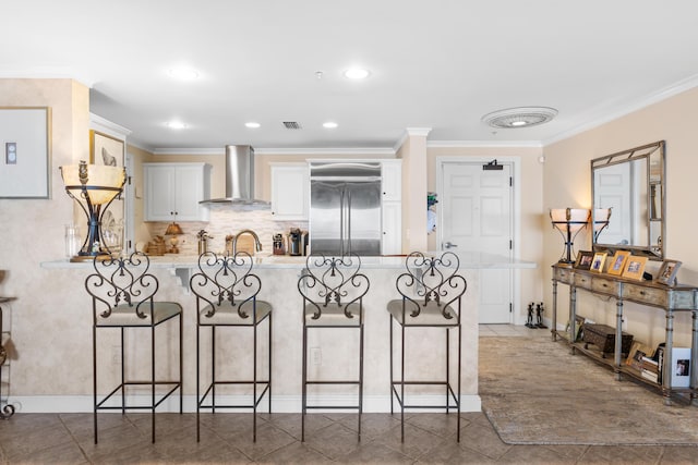 kitchen with white cabinetry, wall chimney exhaust hood, a kitchen breakfast bar, crown molding, and stainless steel built in fridge