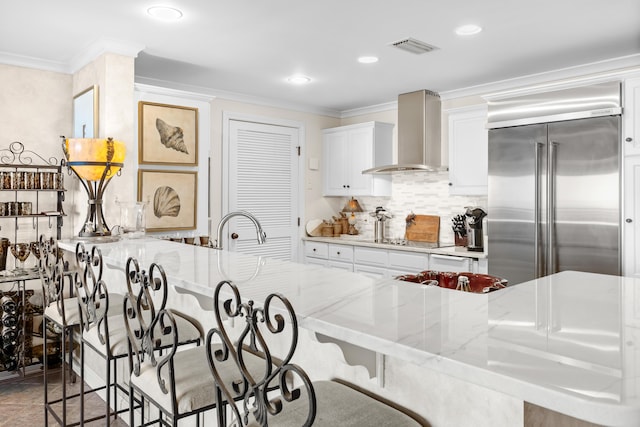 kitchen featuring white cabinetry, stainless steel built in fridge, wall chimney range hood, and light stone counters