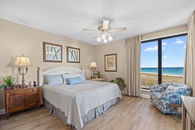 bedroom with ceiling fan, a beach view, light hardwood / wood-style floors, a textured ceiling, and a water view