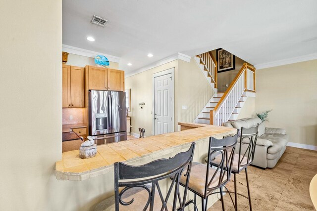 kitchen with a kitchen bar, stainless steel fridge with ice dispenser, tile counters, and crown molding