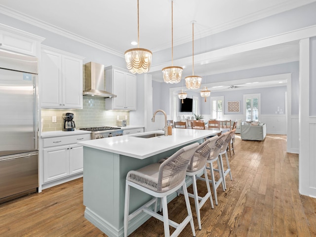 kitchen featuring a kitchen island with sink, white cabinets, wall chimney exhaust hood, decorative light fixtures, and stainless steel appliances