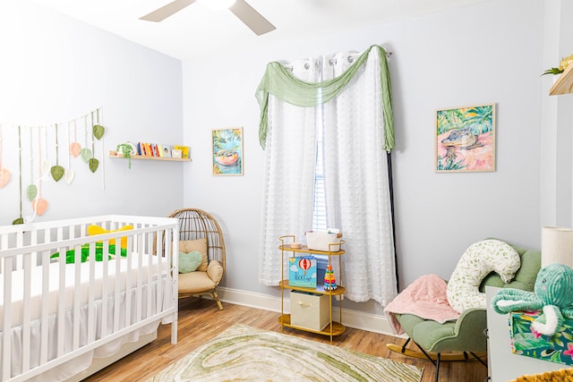 bedroom with a nursery area, a ceiling fan, baseboards, and wood finished floors