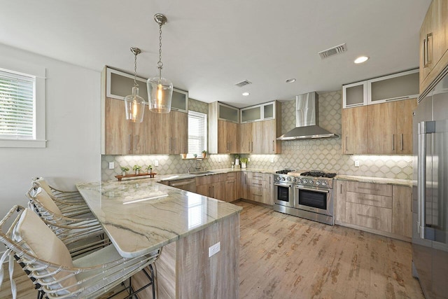 kitchen featuring kitchen peninsula, a kitchen breakfast bar, high end appliances, wall chimney range hood, and pendant lighting