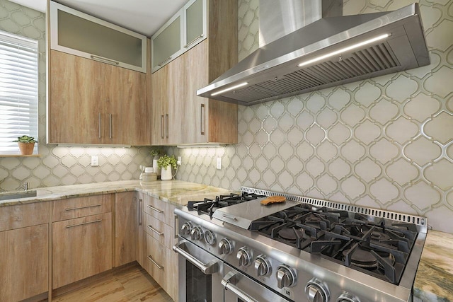 kitchen with decorative backsplash, light stone counters, wall chimney exhaust hood, high end stainless steel range, and light hardwood / wood-style floors