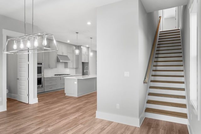 interior space with gray cabinetry, a kitchen island with sink, exhaust hood, hanging light fixtures, and decorative backsplash