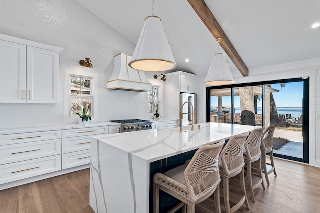 kitchen featuring an island with sink, vaulted ceiling with beams, white cabinets, stove, and custom range hood