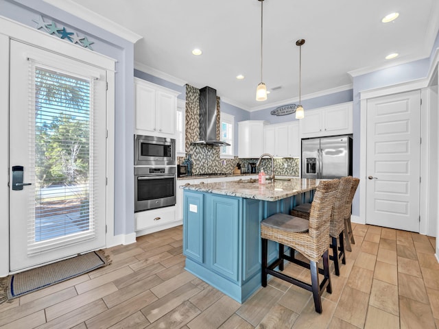 kitchen with light stone countertops, hanging light fixtures, stainless steel appliances, wall chimney range hood, and a kitchen island with sink