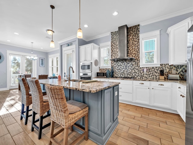 kitchen with white cabinets, wall chimney exhaust hood, light stone countertops, a kitchen island with sink, and appliances with stainless steel finishes