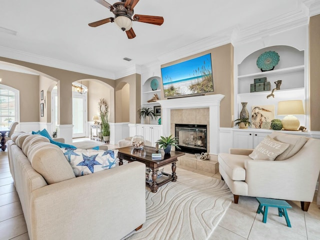 tiled living room featuring ceiling fan with notable chandelier, built in features, ornamental molding, and a tiled fireplace