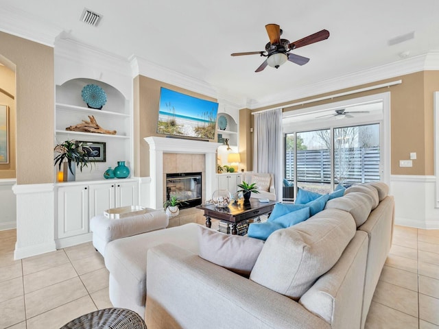 tiled living room featuring built in features, ceiling fan, ornamental molding, and a tiled fireplace