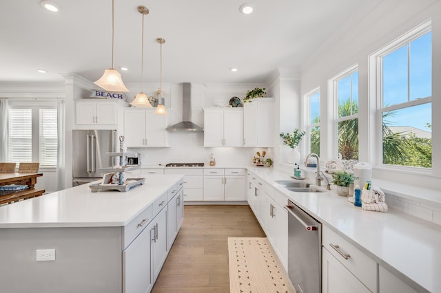 kitchen with appliances with stainless steel finishes, sink, wall chimney range hood, white cabinetry, and hanging light fixtures