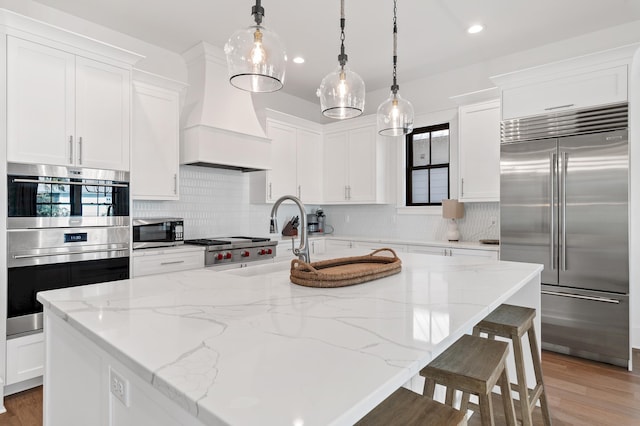 kitchen featuring appliances with stainless steel finishes, custom range hood, a kitchen island with sink, decorative light fixtures, and white cabinetry