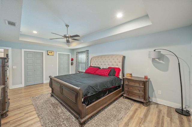 bedroom featuring two closets, light wood-type flooring, a tray ceiling, and ceiling fan