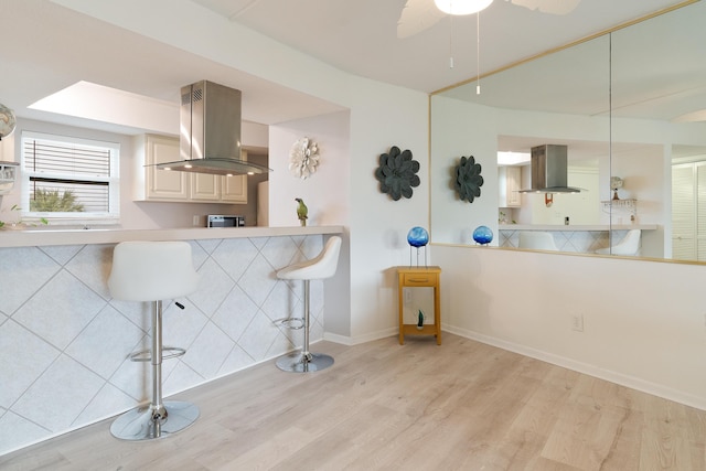 kitchen with light wood-type flooring, a breakfast bar, island range hood, ceiling fan, and wall chimney range hood