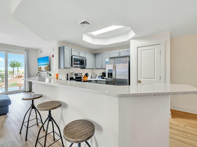 kitchen with gray cabinetry, a kitchen breakfast bar, backsplash, appliances with stainless steel finishes, and light wood-type flooring