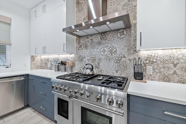 kitchen featuring blue cabinetry, white cabinetry, appliances with stainless steel finishes, decorative backsplash, and wall chimney range hood