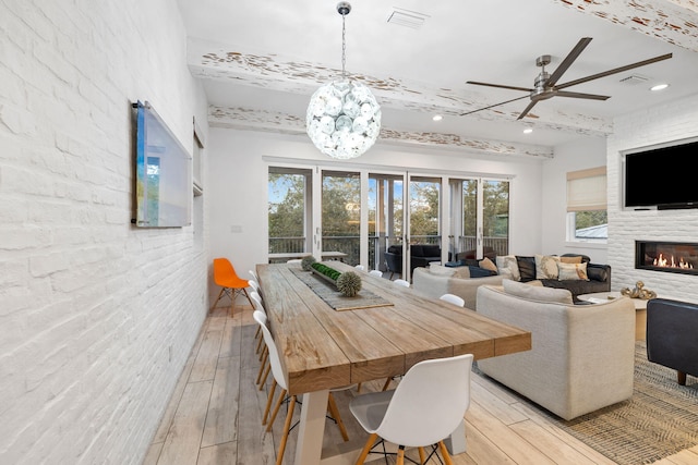 dining room featuring light hardwood / wood-style flooring, beamed ceiling, brick wall, a fireplace, and ceiling fan with notable chandelier