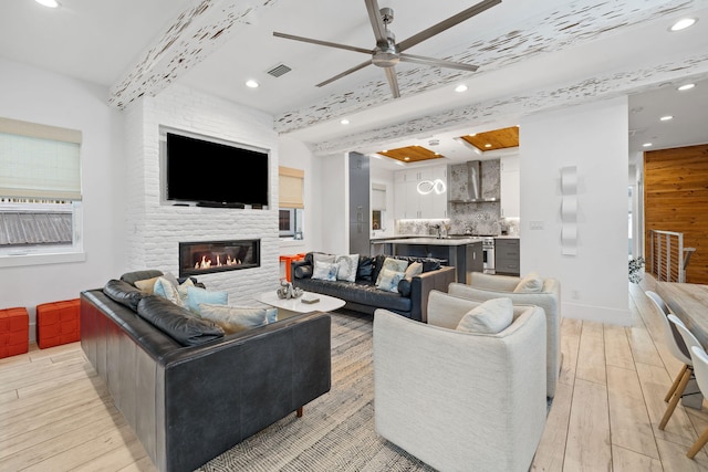 living room featuring beam ceiling, light hardwood / wood-style flooring, a large fireplace, and ceiling fan