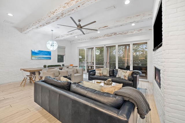 living room featuring brick wall, ceiling fan with notable chandelier, a fireplace, beam ceiling, and light hardwood / wood-style flooring