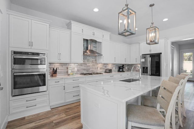 kitchen featuring a kitchen island with sink, white cabinets, sink, wall chimney exhaust hood, and appliances with stainless steel finishes