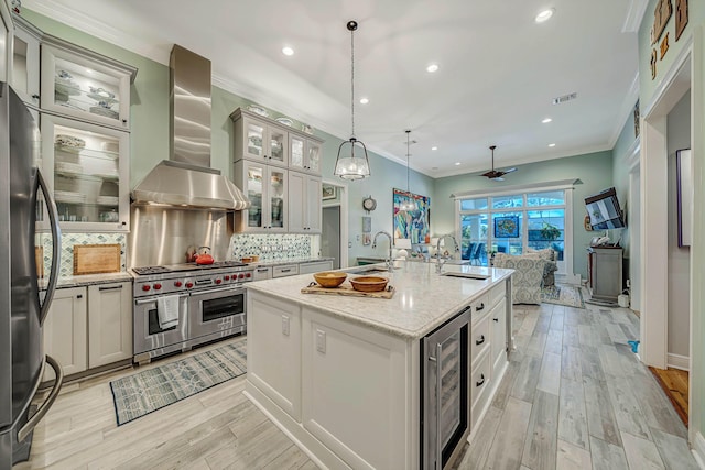 kitchen with sink, wall chimney exhaust hood, decorative light fixtures, a kitchen island with sink, and appliances with stainless steel finishes