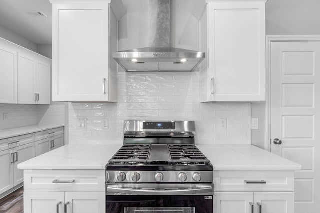 kitchen featuring wall chimney range hood, white cabinetry, light stone counters, stainless steel gas range oven, and decorative backsplash