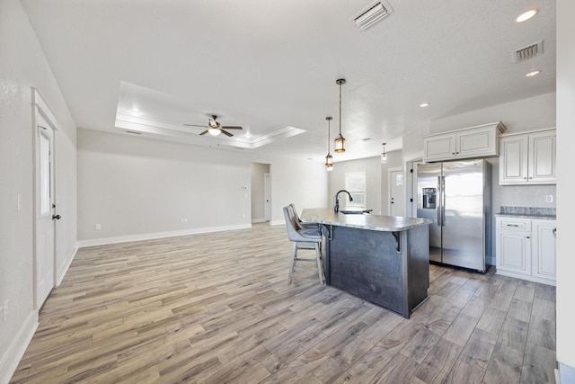 kitchen with ceiling fan, hanging light fixtures, a raised ceiling, stainless steel fridge with ice dispenser, and an island with sink