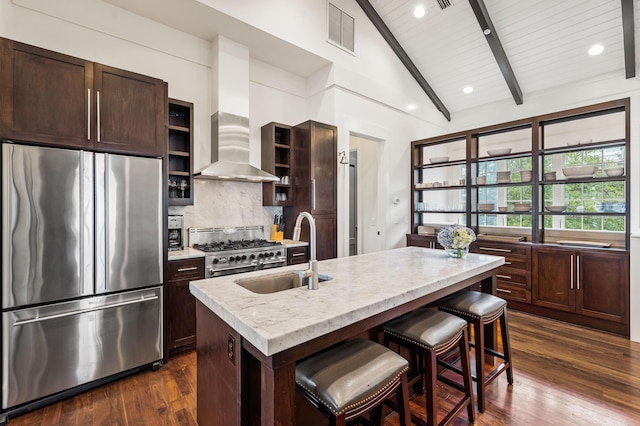 kitchen with sink, wall chimney range hood, an island with sink, a kitchen bar, and appliances with stainless steel finishes