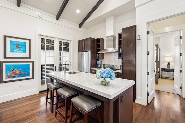 kitchen with sink, wall chimney range hood, lofted ceiling with beams, a center island with sink, and appliances with stainless steel finishes