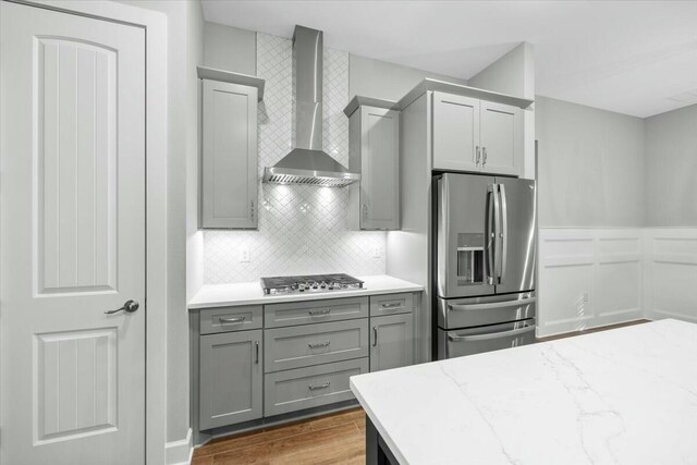 kitchen with wall chimney range hood, gray cabinets, stainless steel appliances, light stone counters, and decorative backsplash