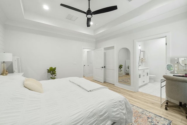 bedroom with a raised ceiling, ensuite bath, ceiling fan, and light hardwood / wood-style flooring