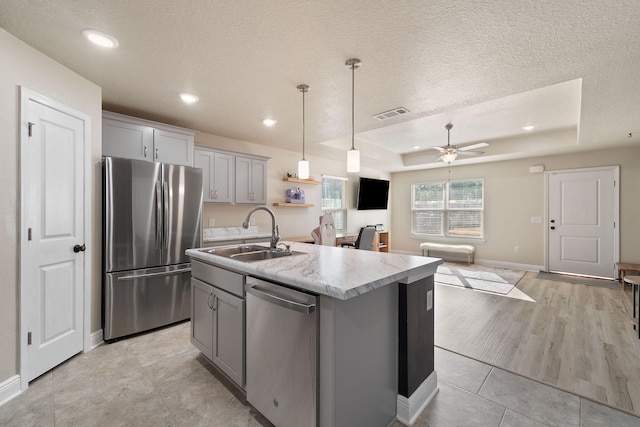 kitchen with gray cabinetry, stainless steel appliances, a raised ceiling, sink, and an island with sink