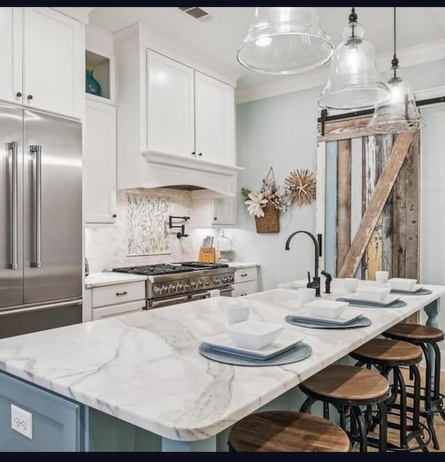 kitchen with light stone counters, high end appliances, decorative light fixtures, a center island with sink, and white cabinetry