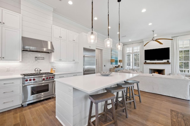 kitchen featuring premium appliances, wall chimney range hood, a fireplace, white cabinetry, and a breakfast bar area