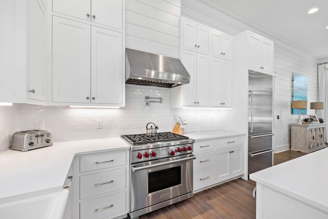 kitchen featuring high end appliances, dark wood-type flooring, white cabinets, wall chimney exhaust hood, and decorative backsplash