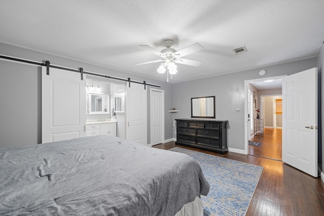 bedroom featuring ensuite bath, a textured ceiling, ceiling fan, dark wood-type flooring, and a barn door