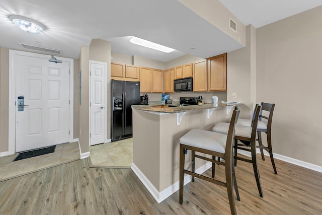 kitchen with light stone countertops, kitchen peninsula, light brown cabinetry, black appliances, and light wood-type flooring