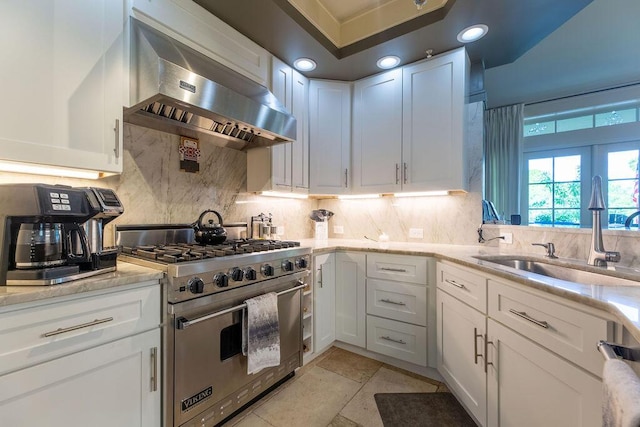 kitchen with luxury stove, sink, wall chimney exhaust hood, light stone counters, and white cabinetry