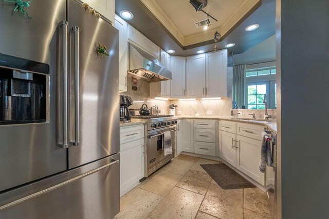 kitchen featuring light stone counters, a raised ceiling, high quality appliances, white cabinetry, and range hood